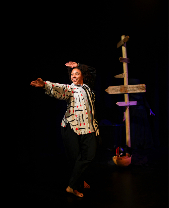 Eshu at the Crossroads - the traveller in a loose multicolour shirt over black pants stands in front of the crossroads sign pointing in her chosen direction