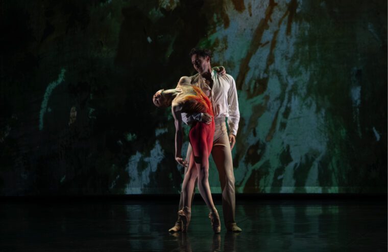 Firebird - A Double Bill. Against a projection of dark, ominous trees, a man in white shirt holds a woman wearing an orange unitard. She's on pointe and leans back over his arm.