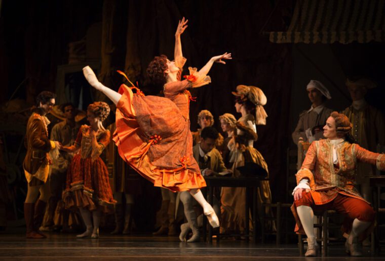 Mayara Magri as Lescaut's Mistress in Manon, dressed in a rich orange costume, leaps in arabesque observed by her patron Monsieur GM, who's sitting to our right