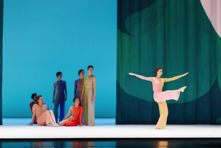 Pastoral, Pam Tanowitz Dance: In front of a green and blue curtain, dancer in a light pink and beige costume stands with one leg raised forward, arms open to the sides, watched by six other dancers