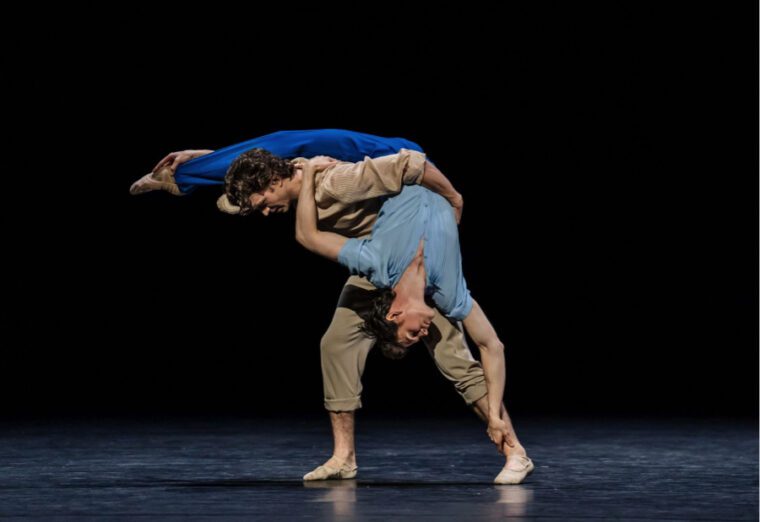 A Centenary Celebration: One male dancer in tan street clothes holes amnother, dressed in blue upside down over his back