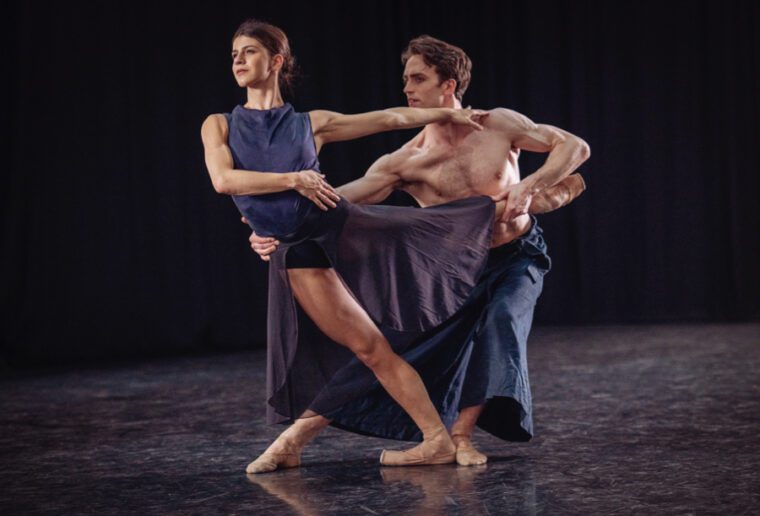 Mayara Magri and Matthew Ball dance a contemporary piece.  They wear indigo costumes. On bent knees he holds her in an off balance arabesque